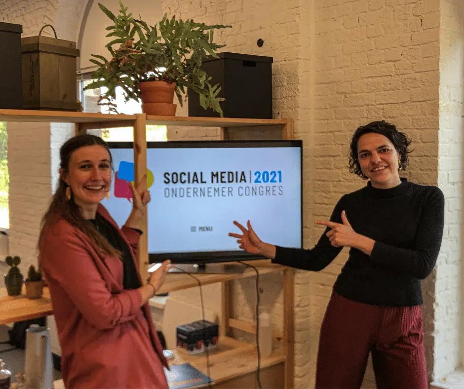 Febe en Ines van Graviteit wijzen enthousiast naar een tv-scherm waarop de tekst "Social Media Ondernemer Congres 2021" staat. De afbeelding suggereert een professionele omgeving en een focus op sociale media.