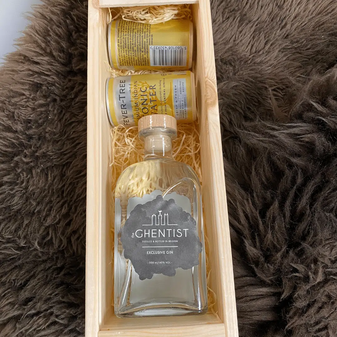 Een houten geschenkdoos met een fles exclusieve gin met het label "Ghentist," vergezeld van twee blikjes tonic water, op een harige achtergrond.