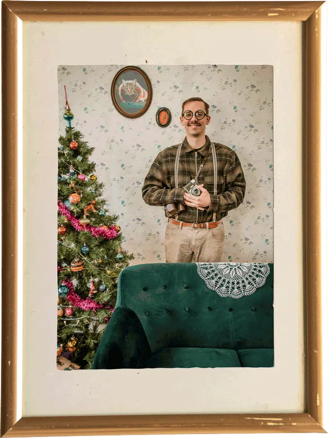 Robbe van Graviteit met bril en snor, gekleed in vintage kleding en met een camera in zijn handen, poseert voor een kerstboom in een ouderwets ingerichte woonkamer, ingelijst in een gouden lijst.