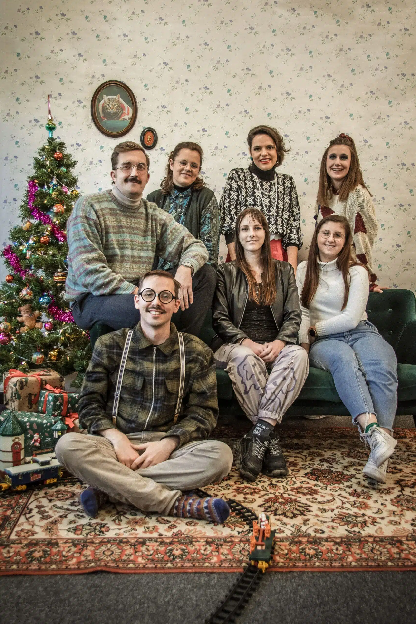 Groepsfoto met kerstthema: Het Graviteit team bestaande uit zeven mensen zit en staat voor een kerstboom in een nostalgische kamer met een tapijt en behang. Ze zijn casual gekleed en lachen naar de camera.