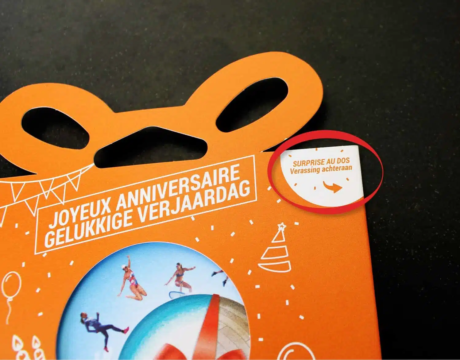 Bovenkant van een oranje cadeaukaart verpakking met handvat en de tekst 'Joyeux Anniversaire' en 'Gelukkige Verjaardag', met een cirkelvormige uitsnede waarin mensen sporten.