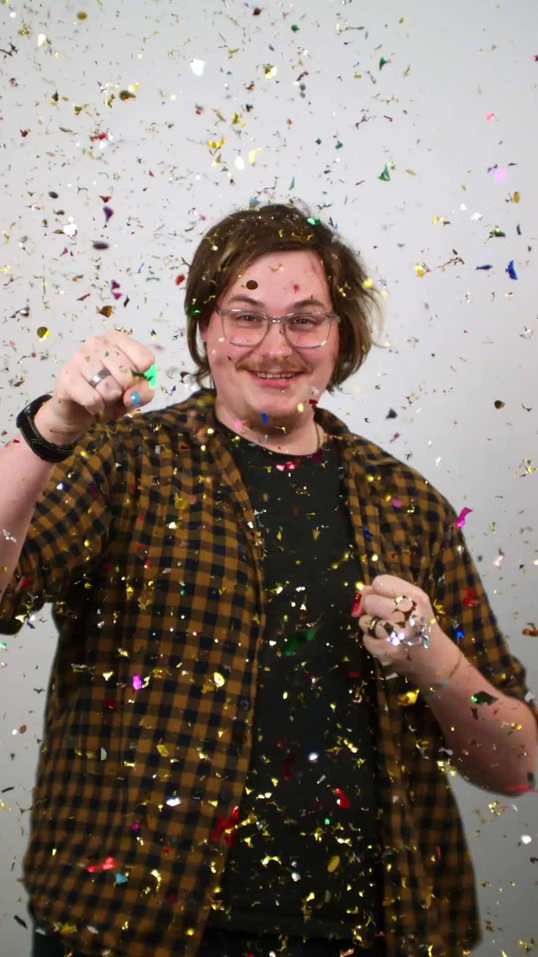 Een man met halflang haar en een bril, gekleed in een geruit overhemd, staat te vieren met confetti om hem heen. Hij glimlacht breed.
