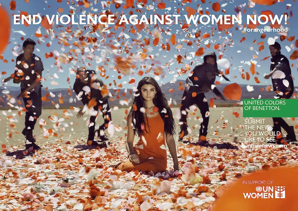 United Colors of Benetton anti-geweld campagne: Afbeelding van een vrouw in een oranje jurk omringd door vallende oranje bloemblaadjes. Op de achtergrond zijn mannen te zien die de bloemblaadjes gooien. De tekst roept op om geweld tegen vrouwen te stoppen.