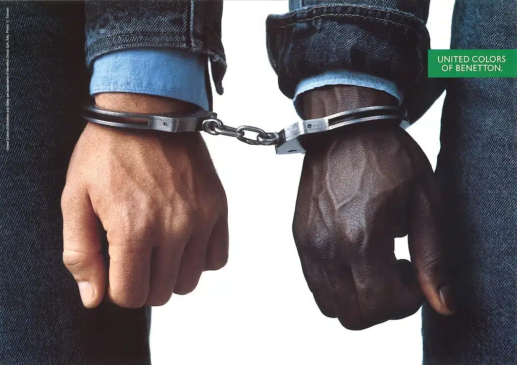 United Colors of Benetton anti-discriminatie campagne: Close-up van twee mannenhanden, één met een lichte huidskleur en één met een donkere huidskleur, vastgebonden met handboeien. De afbeelding symboliseert verbondenheid en anti-discriminatie.
