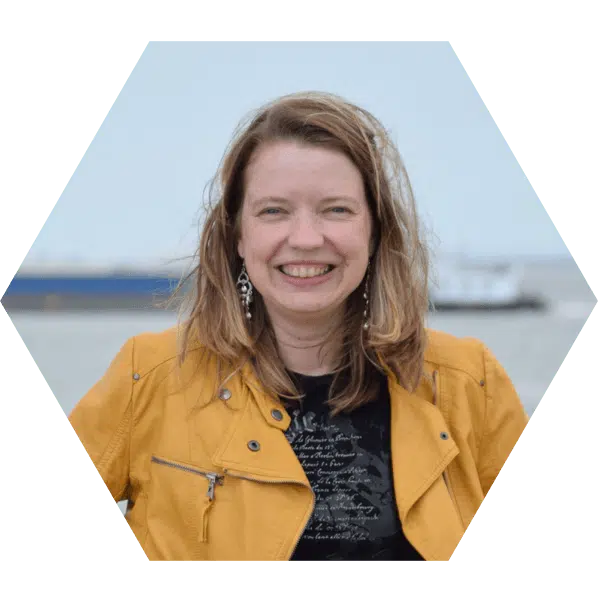 social media ondernemer congres Een hexagonale foto van een vrouw met halflang blond haar, gekleed in een gele jas. Ze staat buiten met een waterrijke achtergrond en glimlacht breed naar de camera.