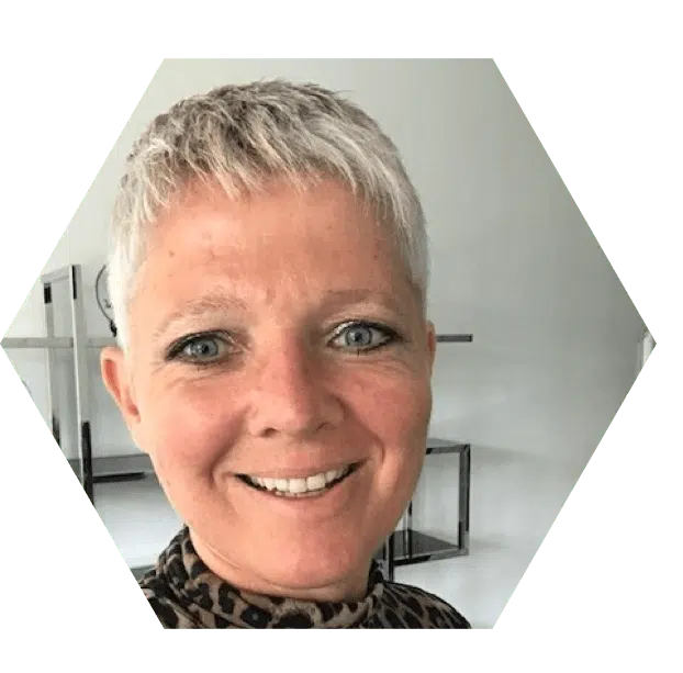 social media ondernemer congres Een hexagonale foto van een vrouw met kort, grijs haar en een breed glimlachende uitdrukking. Ze draagt een blouse met een patroon en bevindt zich in een lichte kamer met een moderne uitstraling.