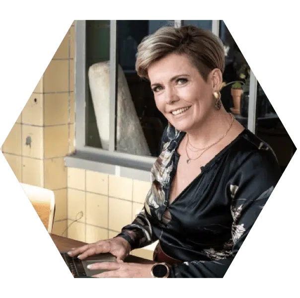 social media ondernemer congres Een hexagonale foto van een vrouw met kort blond haar, gekleed in een donkerblauwe blouse. Ze zit aan een bureau en typt op een laptop, glimlachend naar de camera. De setting lijkt een lichte en moderne werkruimte.