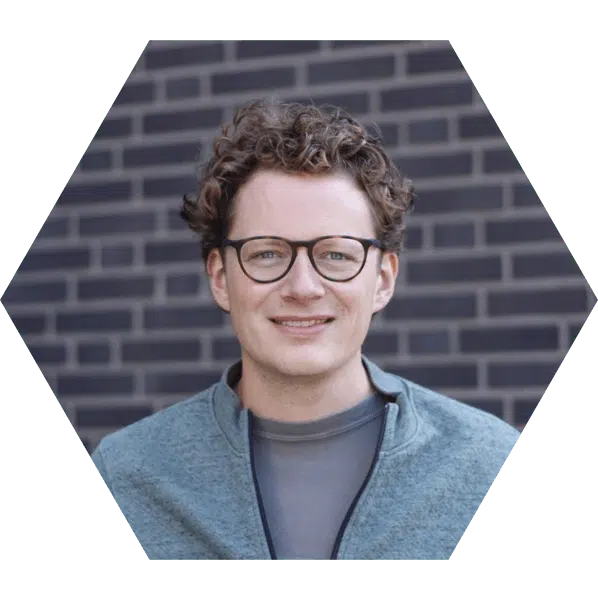 social media ondernemer congres Een hexagonale foto van een man met krullend haar en een bril. Hij draagt een grijze trui en staat voor een donkergrijze muur, glimlachend naar de camera.