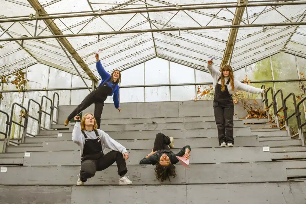 Het creatieve team van Graviteit poseert speels op betonnen tribunes in een verlaten serre met papieren vliegtuigjes in de ha