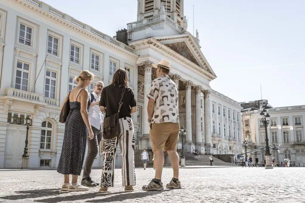 Vier mensen in gesprek op een zonnig plein voor de neoclassieke gevel van de Koningsplaats in Brussel tijdens een Karel Guide